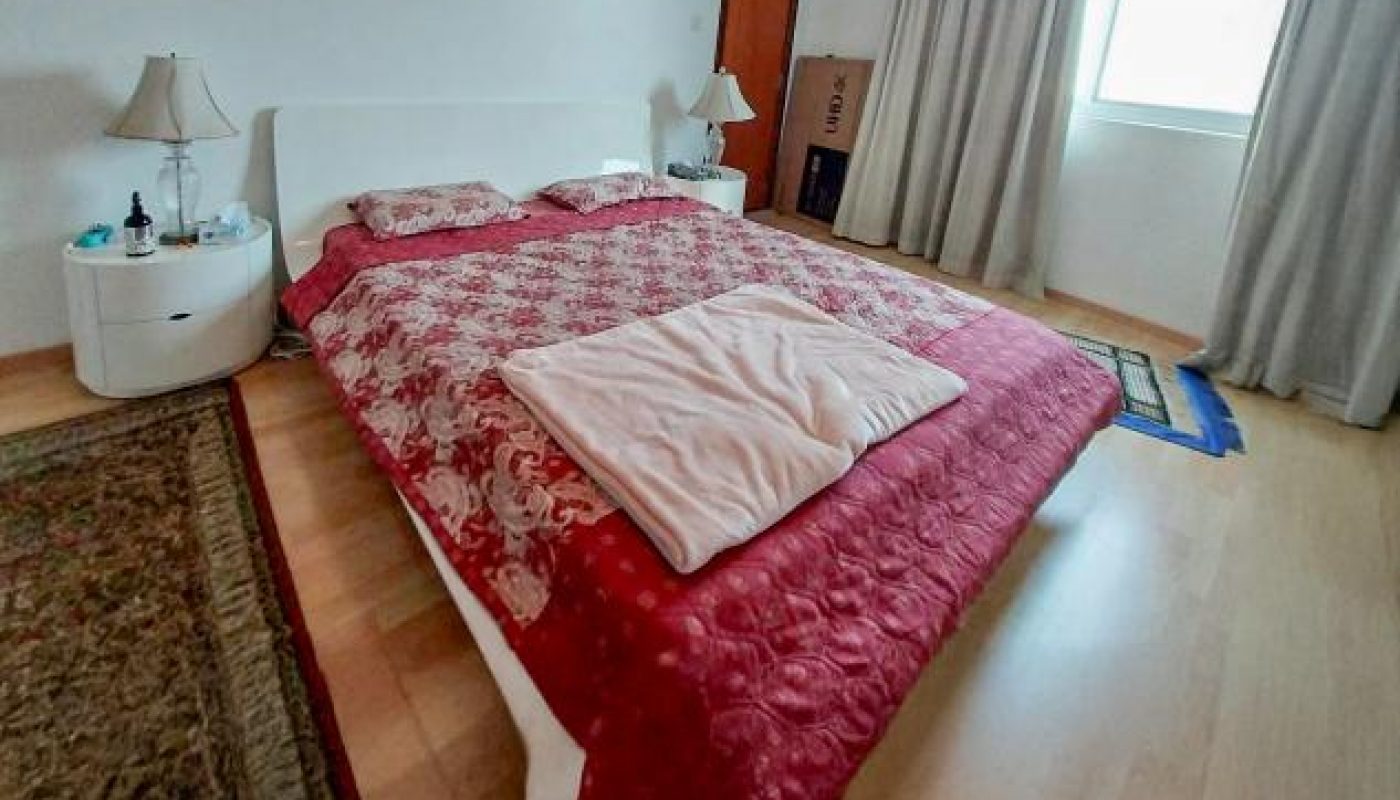 A bedroom with a double bed covered in a red patterned blanket, two bedside tables with lamps, a window with curtains, and a rug on light wood flooring.