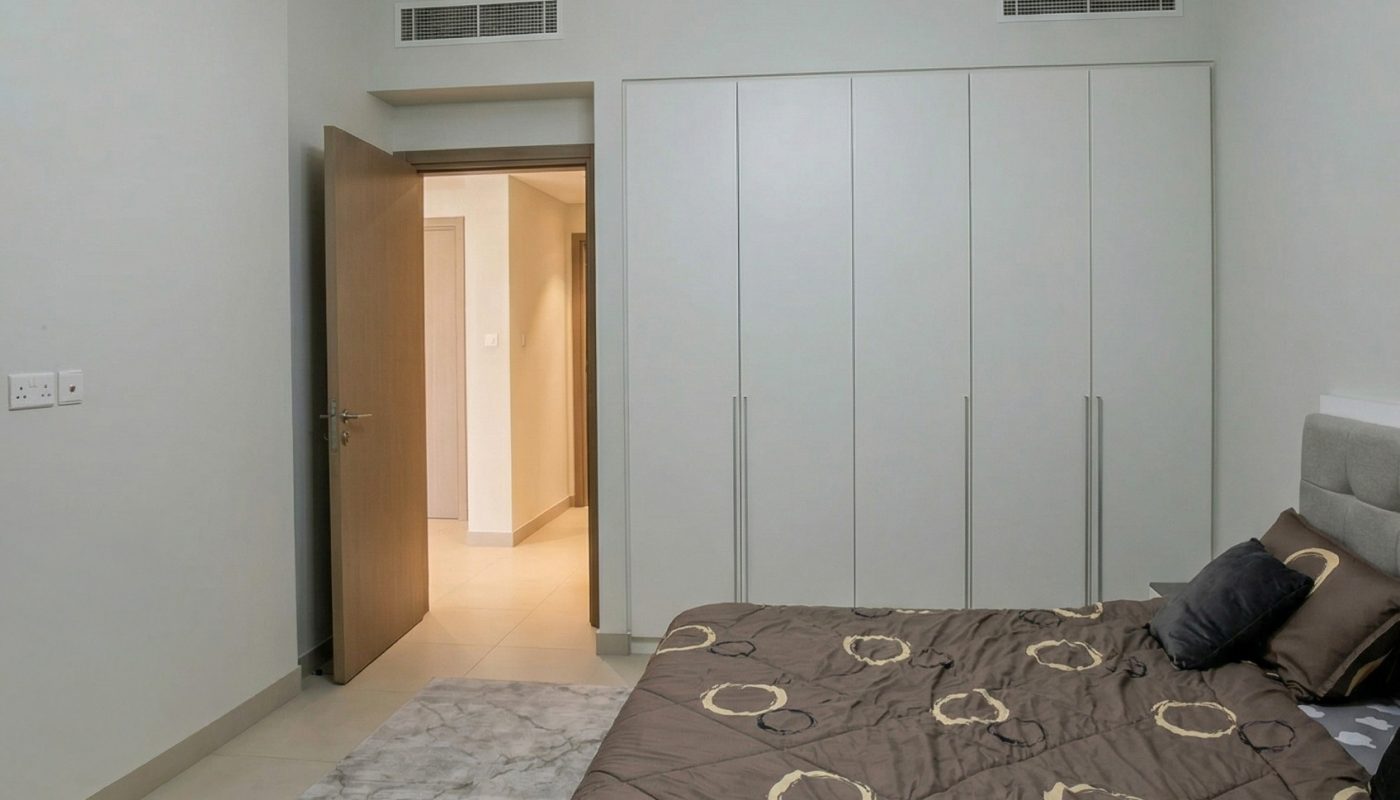 Modern bedroom with a double bed, brown patterned bedding, a gray rug, built-in white wardrobes, an open door, and a ceiling light fixture with round bulbs.