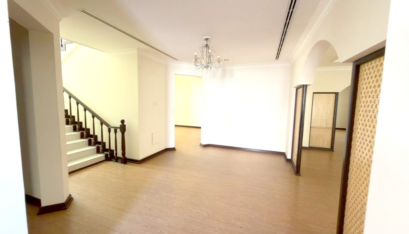 Spacious empty room with light wooden flooring, white walls, a staircase with dark railing, a chandelier, and arched doorways.
