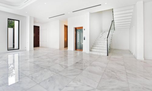 A spacious, modern white interior with glossy marble floors, white walls, a staircase with a glass railing, large windows, and a wooden door characterizes this luxury villa.