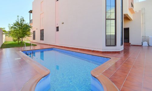 A narrow, curved swimming pool with red-tiled flooring lies in the backyard of a modern 3BR villa. The pool is surrounded by a beige border, adjacent to the stylish two-story building, with a tree standing proudly in the background amidst the big garden.