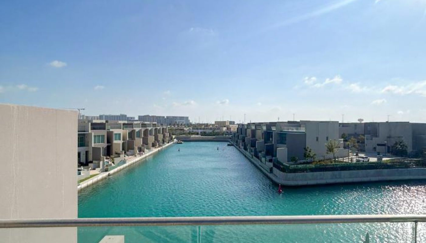 Rows of modern waterfront houses line both sides of a turquoise canal under a bright blue sky, seen from a balcony with a glass railing.