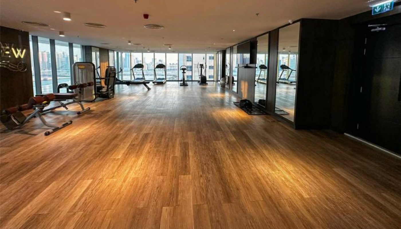 Spacious gym with wooden flooring, exercise equipment along the windows, and city views through large glass panels.