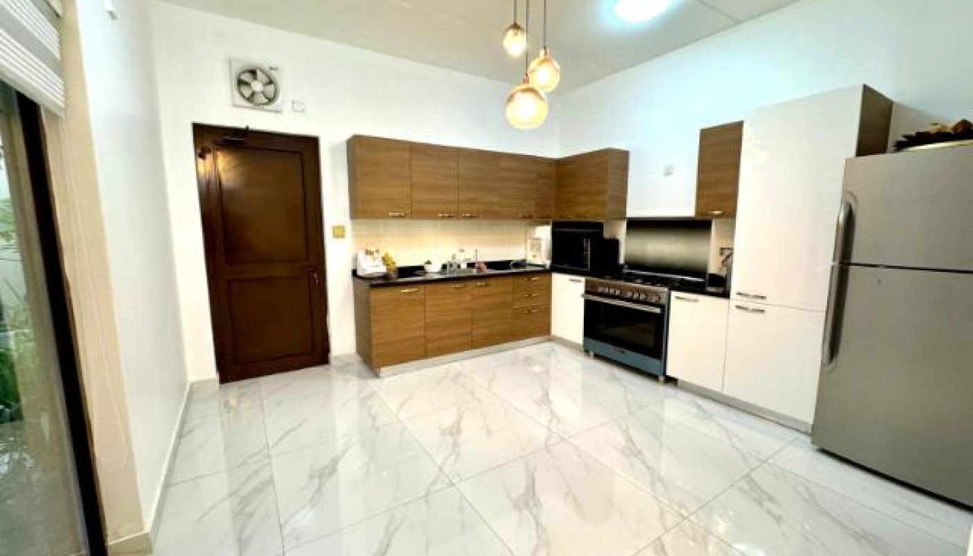 Modern kitchen with wood cabinets, white countertops, stainless steel appliances, tiled floor, and pendant lights. Sliding glass door and brown wooden door are visible.