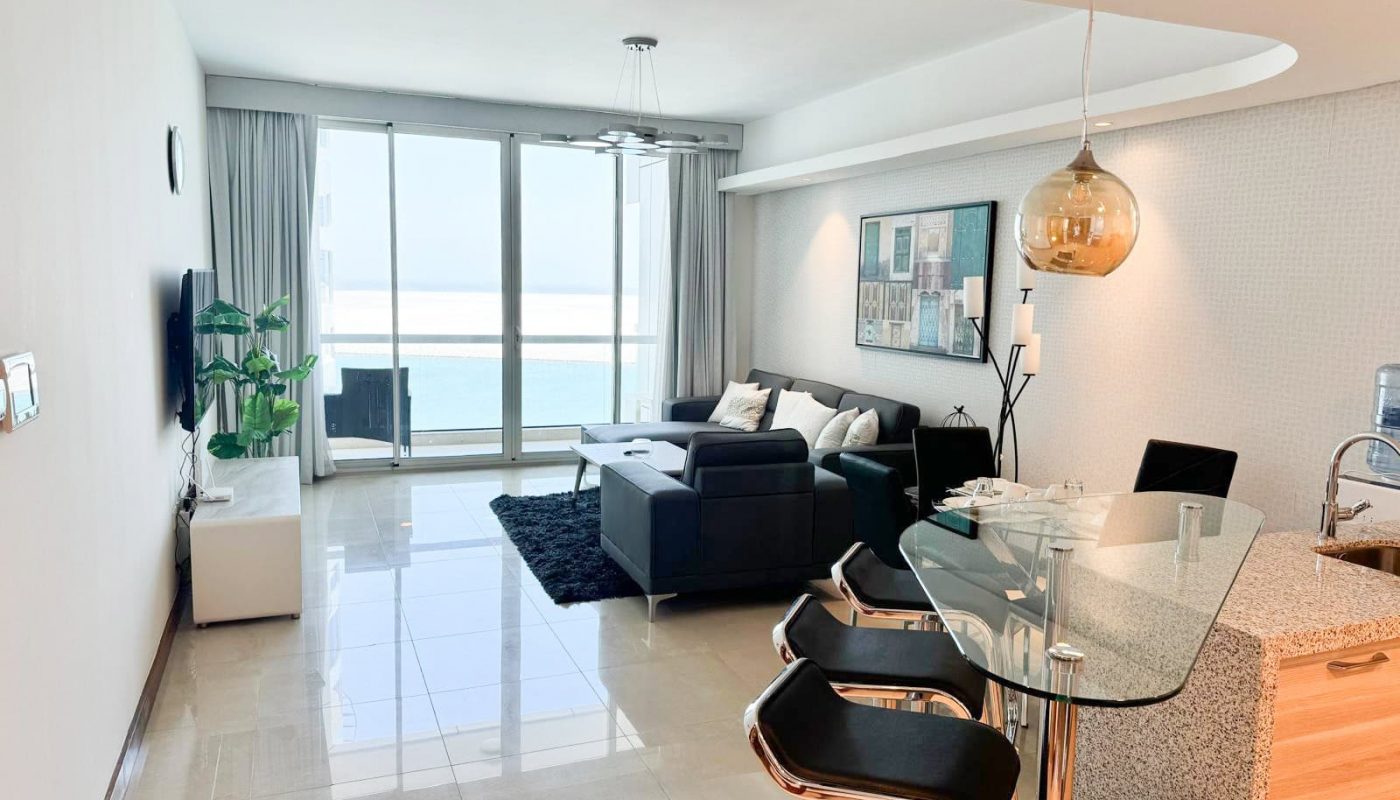Modern living room and kitchen area with large windows overlooking the ocean, a black sectional sofa, glass dining table with barstools, and bright natural light.