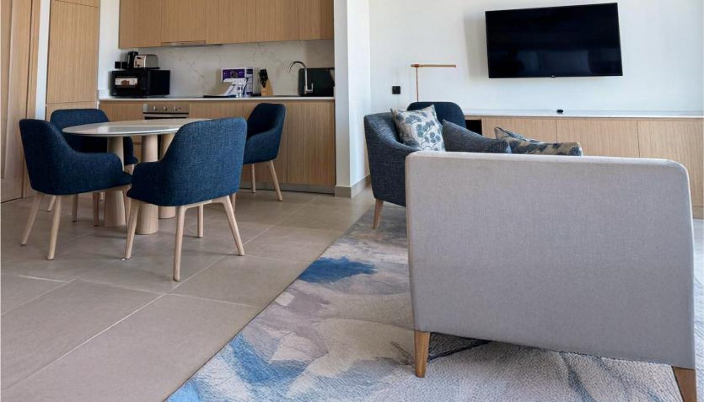 Modern open-plan living area with a small kitchen, dining table with four blue chairs, a seating area with armchairs, wall-mounted TV, and a rug on the tiled floor.