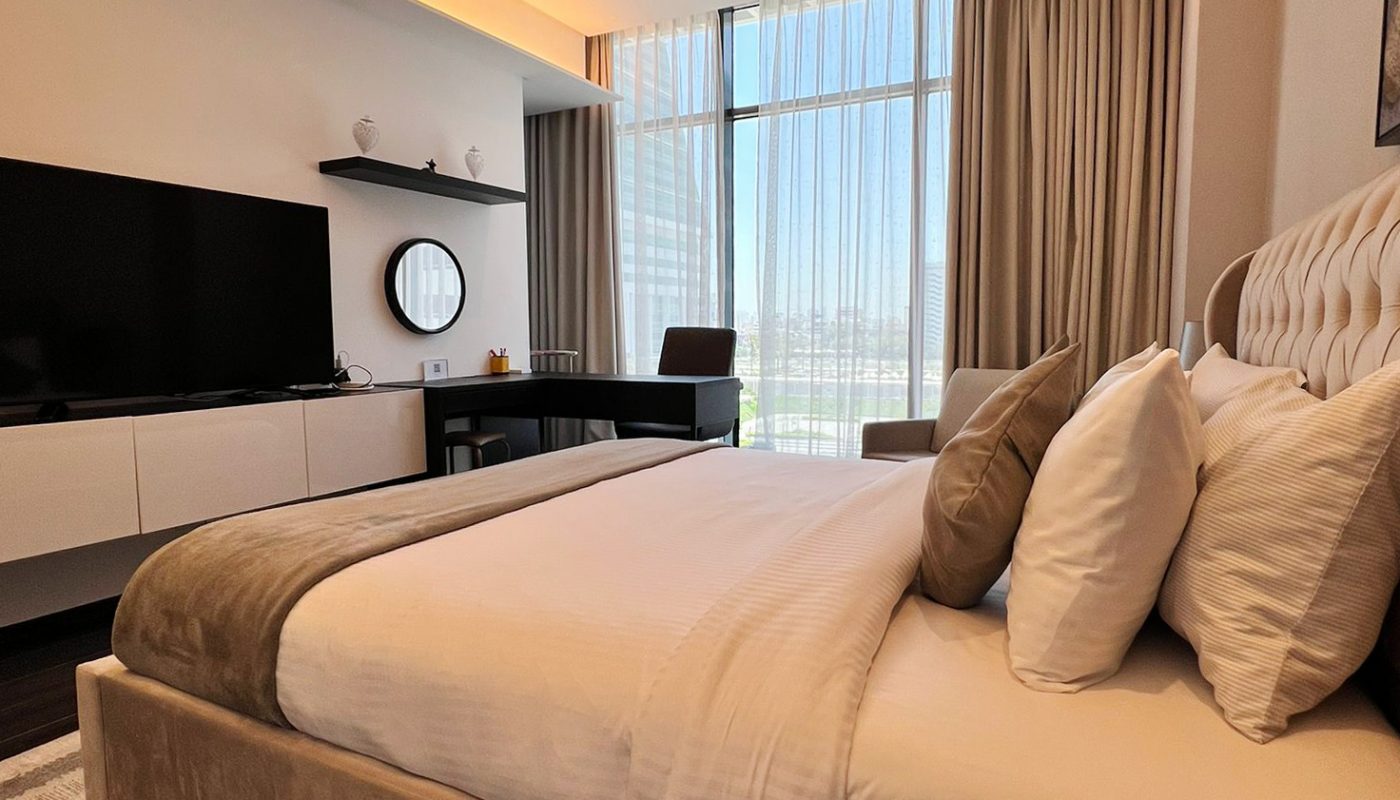 Modern hotel room with a large bed, neutral decor, flat-screen TV, desk, large window with sheer curtains, and a city view.