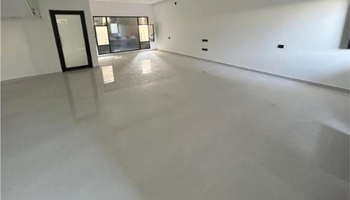 Empty room with white walls, glossy floor, ceiling lights, a door to the left, and large windows at the far end letting in natural light.