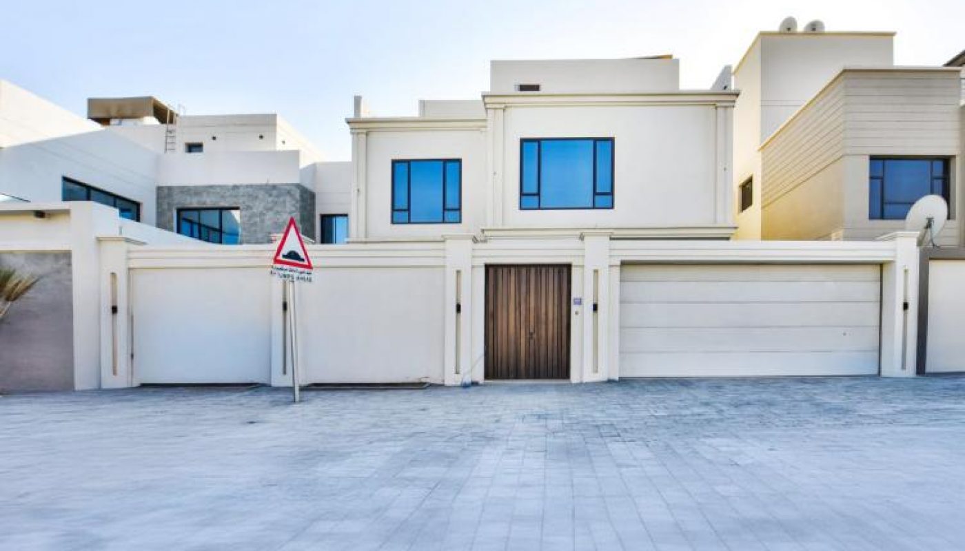 Modern two-story house with large windows, white exterior walls, a wooden front door, and a closed garage. A traffic sign is visible in front of the property.