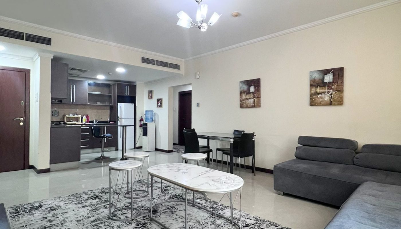 Modern living room with a gray sectional sofa, abstract rug, coffee tables, dining table with four chairs, and an open view into the kitchen area. Neutral tones and wall art are present.