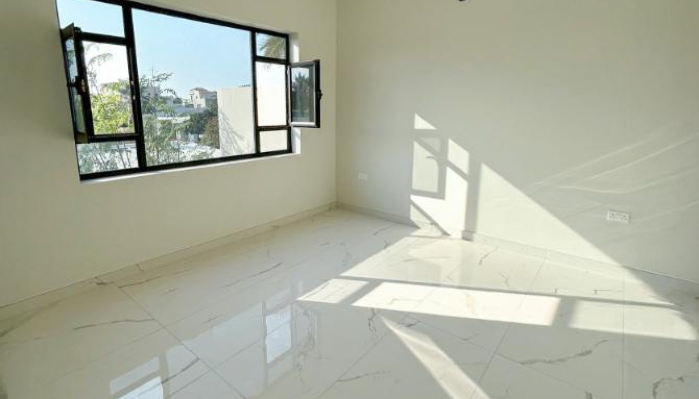Empty room with large window, white walls, and glossy white marble floor tiles. Sunlight casts shadows on the floor, and recessed ceiling lights are visible.