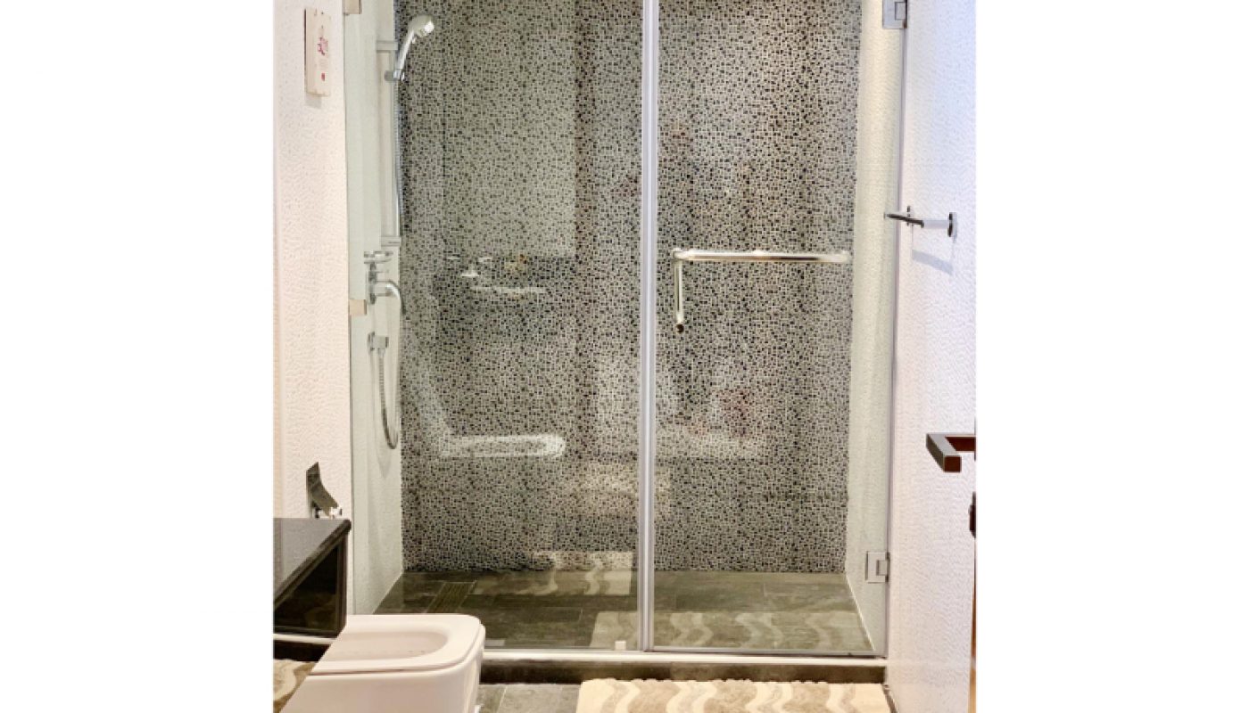 Modern bathroom with a glass shower enclosure, handheld showerhead, toilet, black countertop, and patterned bath mats on a tiled floor.