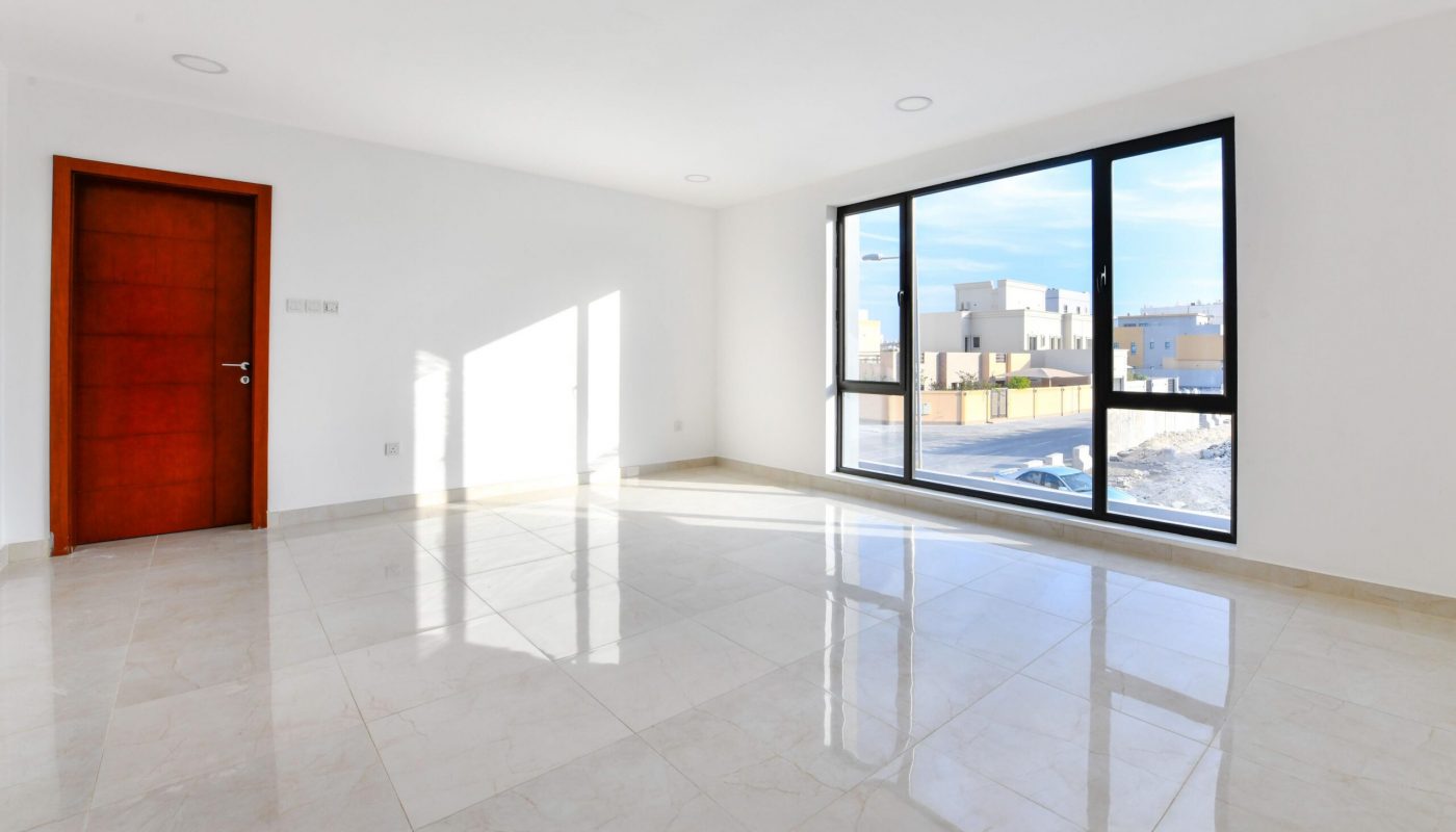 Empty, sunlit room with white walls, large floor tiles, a wooden door on the left, and a wide window showing a neighborhood street outside.
