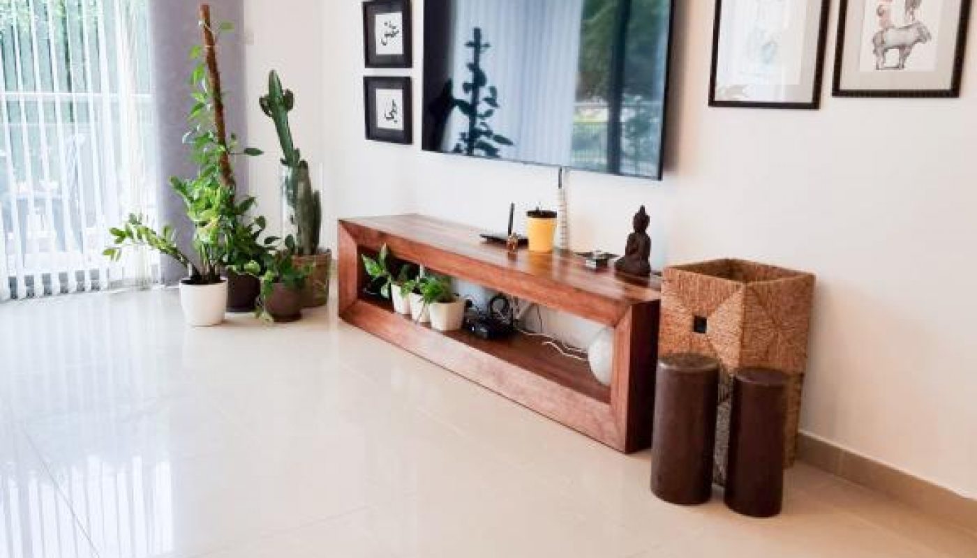 Modern living room with a wall-mounted TV, wooden console, potted plants, framed artwork, and woven baskets on a shiny tiled floor.