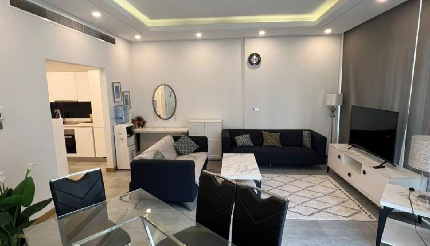 Modern living and dining area with glass dining table, black chairs, navy sofas, wall-mounted TV, and kitchen visible in the background. Light gray floor and recessed ceiling lighting.