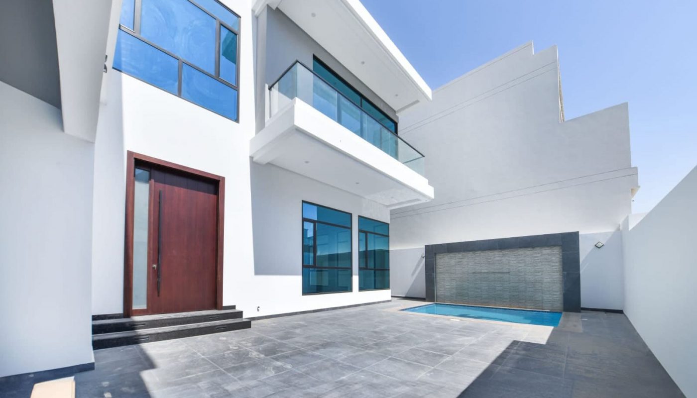 Modern two-story white building with large windows, a wooden door, and a balcony. There is an adjacent swimming pool and paved courtyard under a clear blue sky.