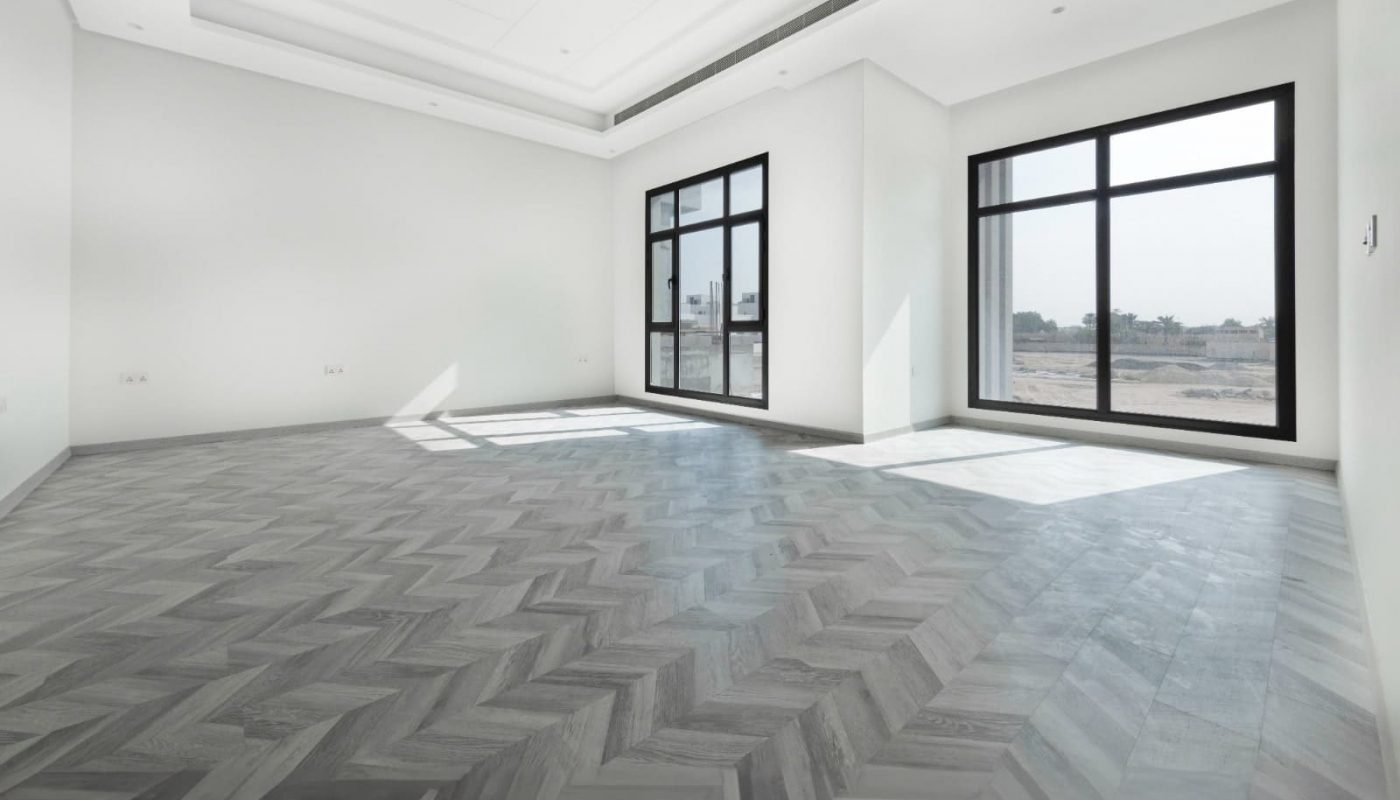 Spacious empty modern room with herringbone-patterned flooring, large windows, and white walls.
