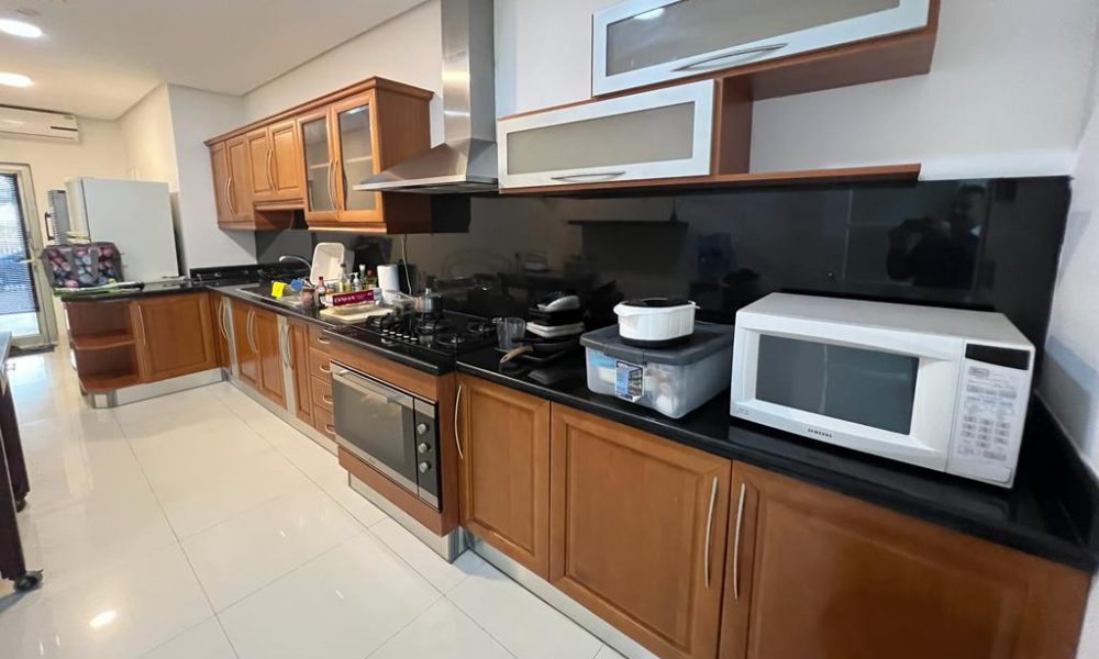 Spacious kitchen with wooden cabinets, black countertops, a stove, microwave, and various appliances on the counter. White tiled floor and wide hallway.