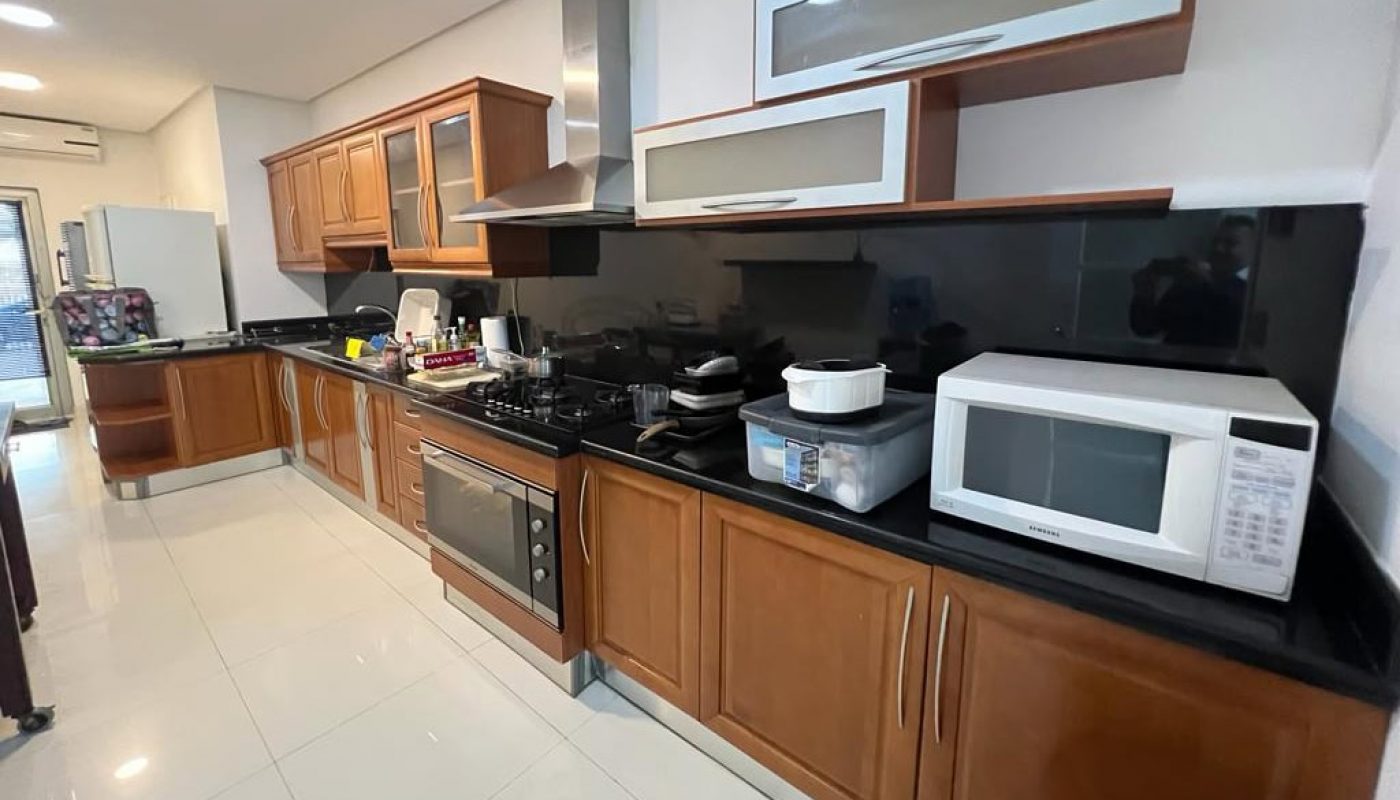 Spacious kitchen with wooden cabinets, black countertops, a stove, microwave, and various appliances on the counter. White tiled floor and wide hallway.
