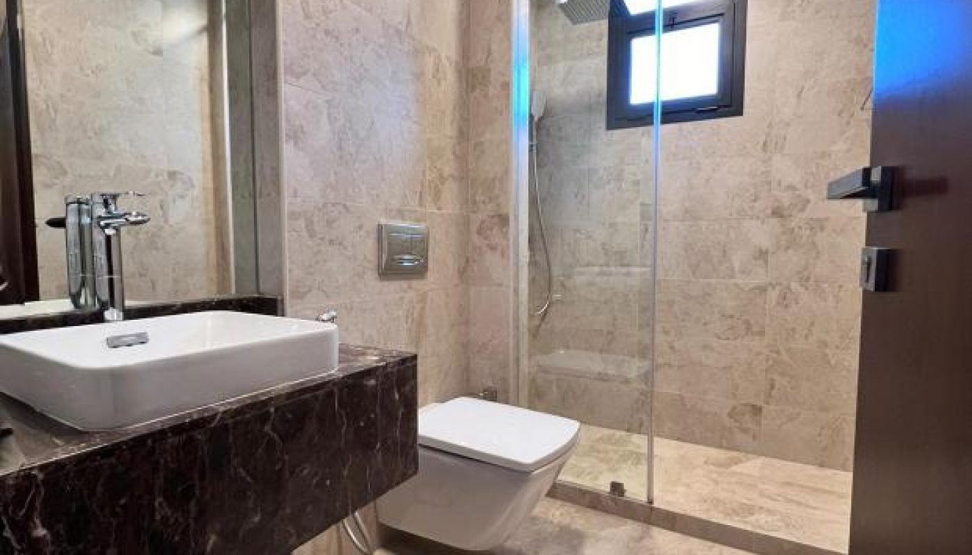 Modern bathroom with beige marble tiles, a wall-mounted toilet, vessel sink on a dark vanity, and a glass-enclosed shower with a window above.