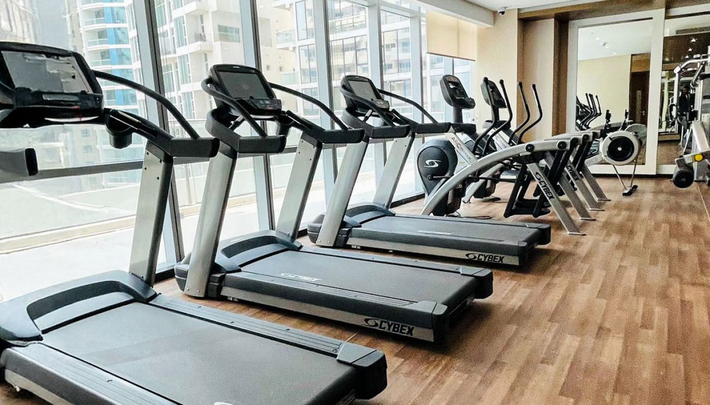 A modern gym with several treadmills and exercise machines lined up on a wooden floor near large windows.