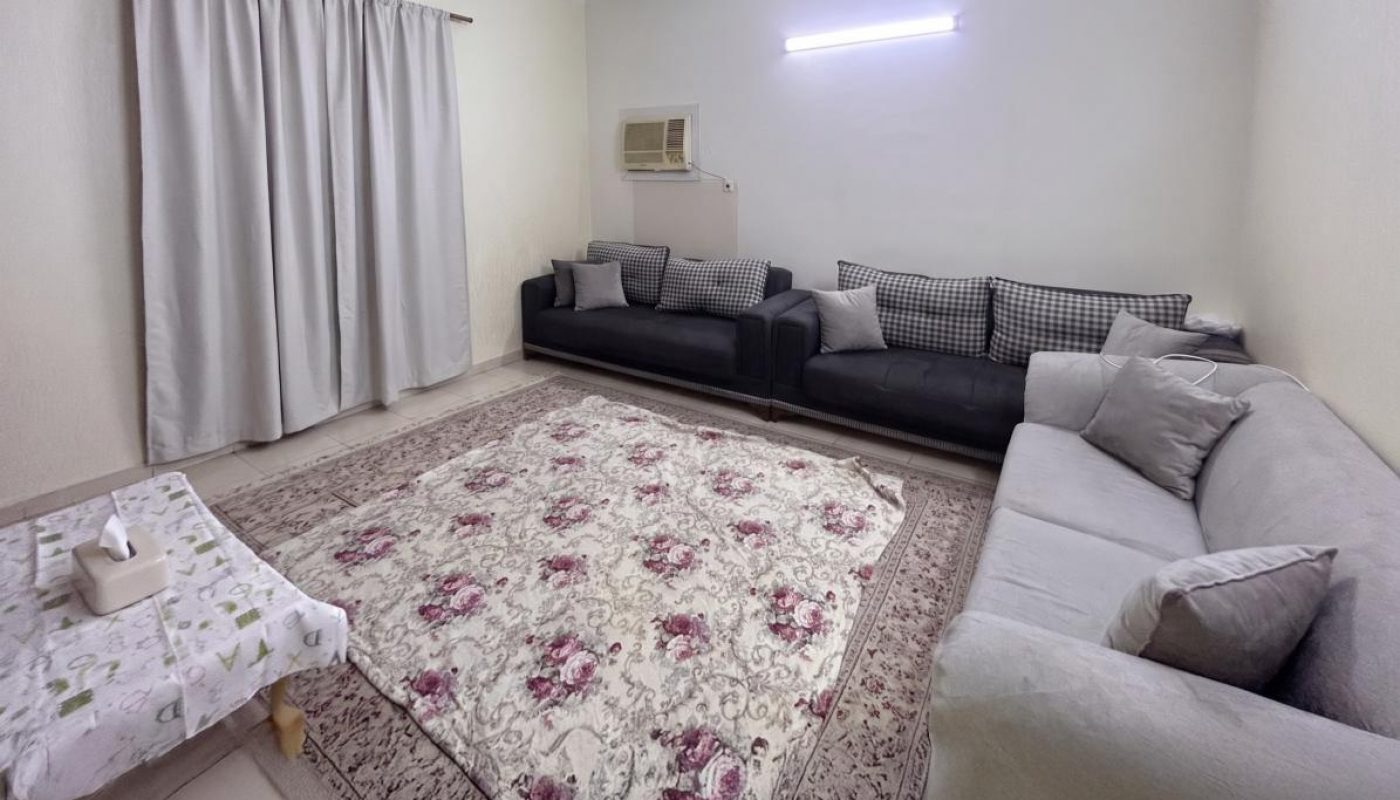 A living room with two sofas, a ceiling fan, an air conditioner, a patterned rug, a coffee table with a tissue box, and closed curtains on one wall.
