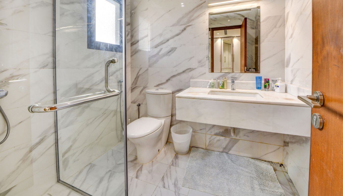 Modern bathroom with marble walls and floor, glass shower enclosure, toilet, sink with counter, mirror, and a small window. A gray rug lies on the floor in front of the sink.