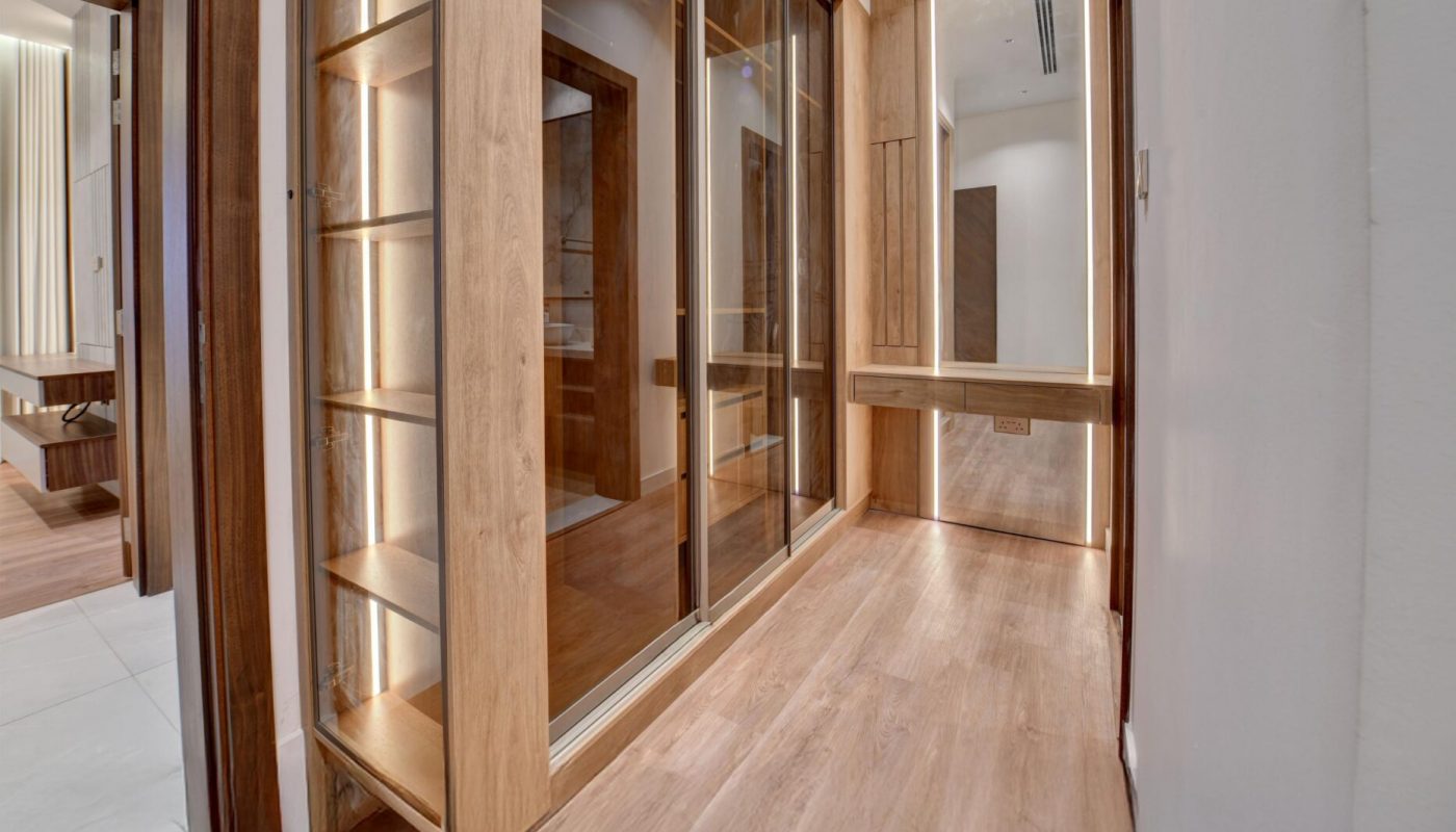Modern walk-in closet with wooden floors, built-in shelves, glass sliding doors, and overhead lighting, viewed from the hallway entrance.