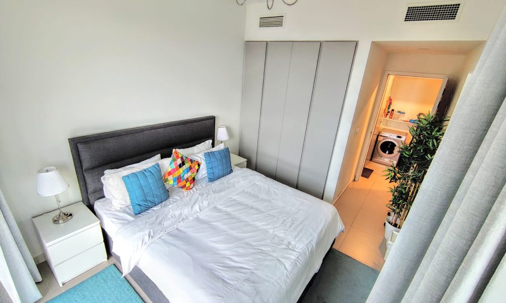 A bedroom with a neatly made double bed, colorful cushions, and bedside lamps. There is a closet and an open door leading to a room with a washer and dryer.