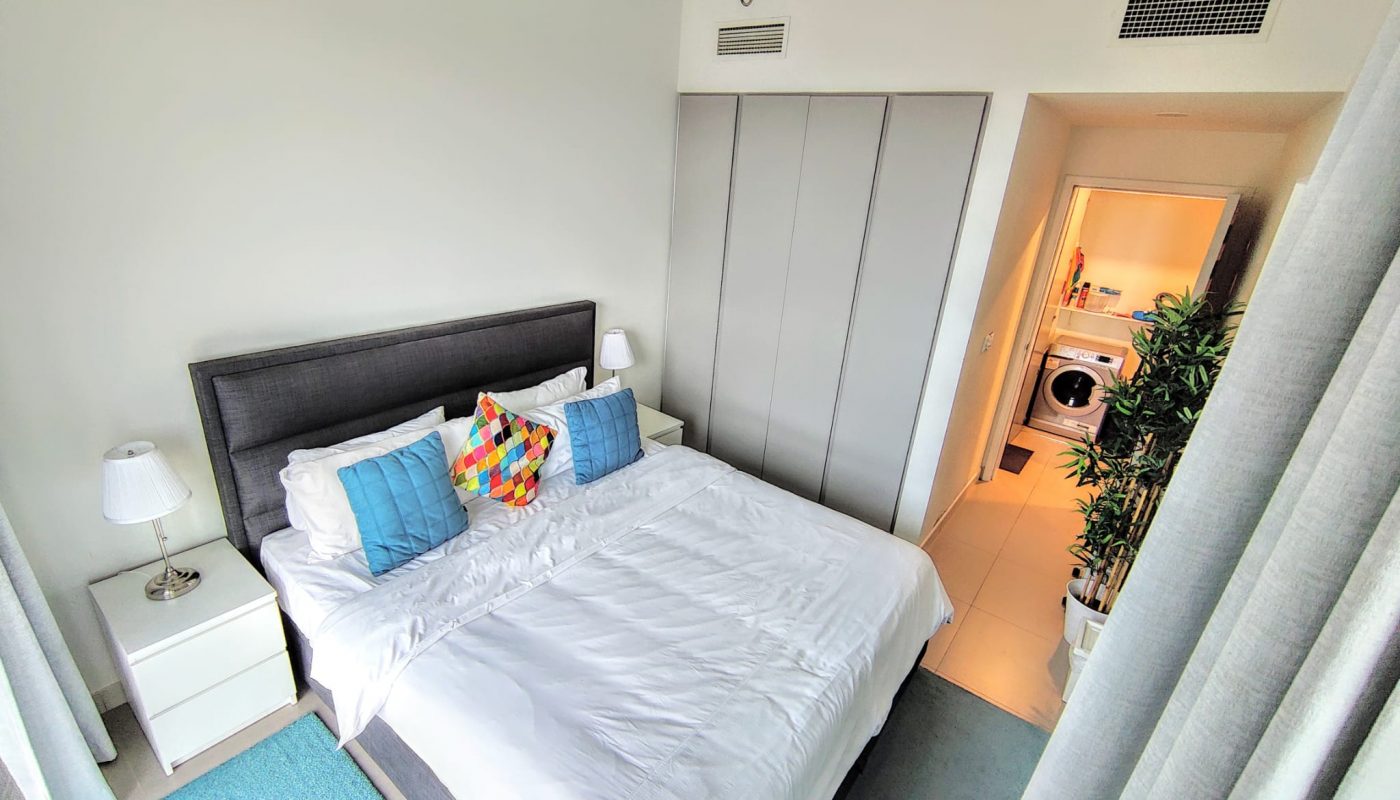 Bedroom with a large bed, three colorful pillows, two bedside tables with lamps, wardrobe, and view into a laundry area with a washing machine and dryer.