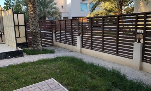 A pathway lined with grass and palm trees leads to a modern building, separated by a wooden fence with horizontal slats.