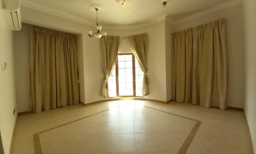 A spacious, empty room with cream-colored curtains, a tiled floor, a ceiling fan, and overhead light fixtures. Natural light enters through a large window.