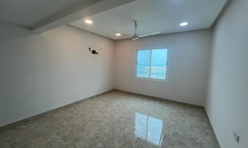 An empty room in an available apartment for rent, located in the Galali area, with a tiled floor, a ceiling fan, white walls, and a large window with a view outside.