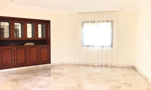 A spacious, empty room with a tiled floor, a large window with sheer curtains, and a dark wood built-in display cabinet on the left wall.