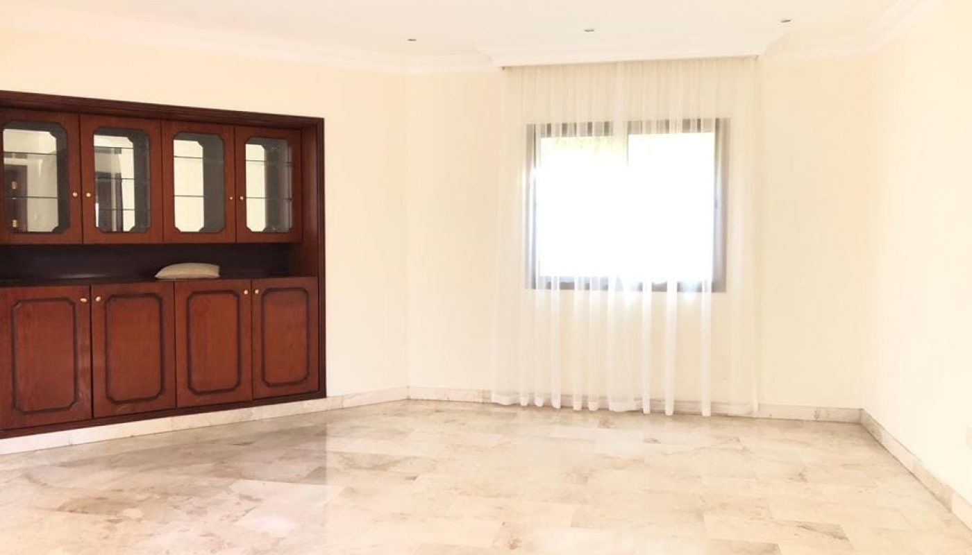 A spacious, empty room with a tiled floor, a large window with sheer curtains, and a dark wood built-in display cabinet on the left wall.