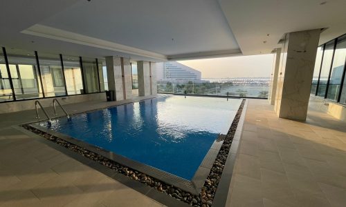 Indoor swimming pool at AMWAJ surrounded by large windows with a view of an outside building and landscape in the background. Stones decorate the pool's border, and pool steps are visible on one side. Ideal for those seeking a luxurious flat for sale.