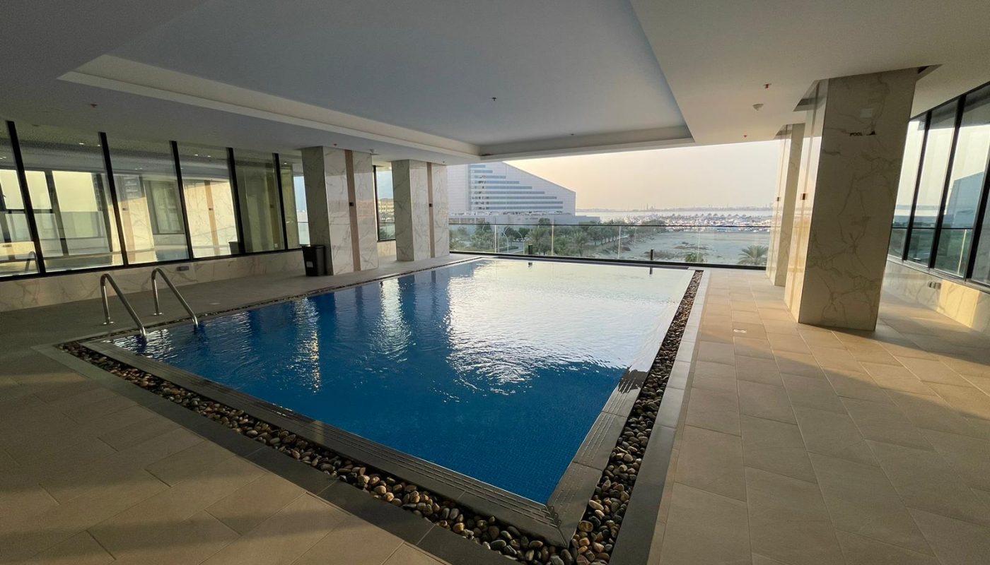 Indoor swimming pool with stone border, surrounded by large windows offering views of a modern building and the outdoor landscape.