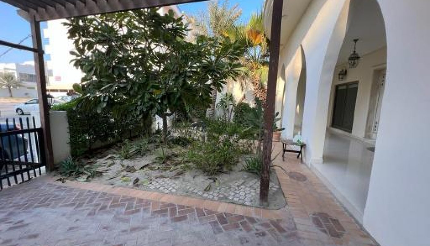 Outdoor patio area with tiled flooring and a garden under a wooden pergola. Arched walkway on the right, and a street visible in the background.