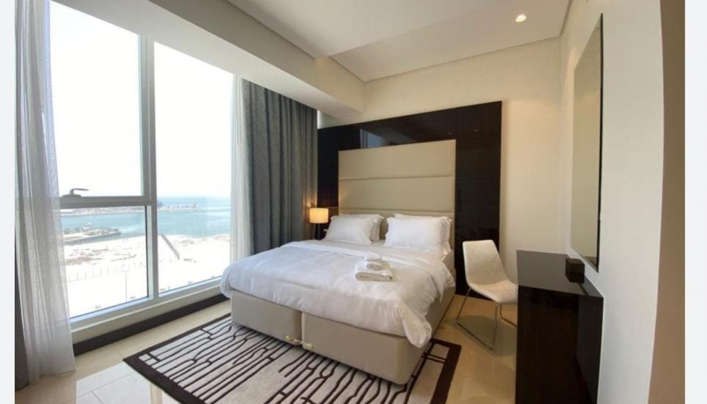 A modern hotel room with a large bed, a chair, a small desk, and a floor-to-ceiling window offering a view of the sea.