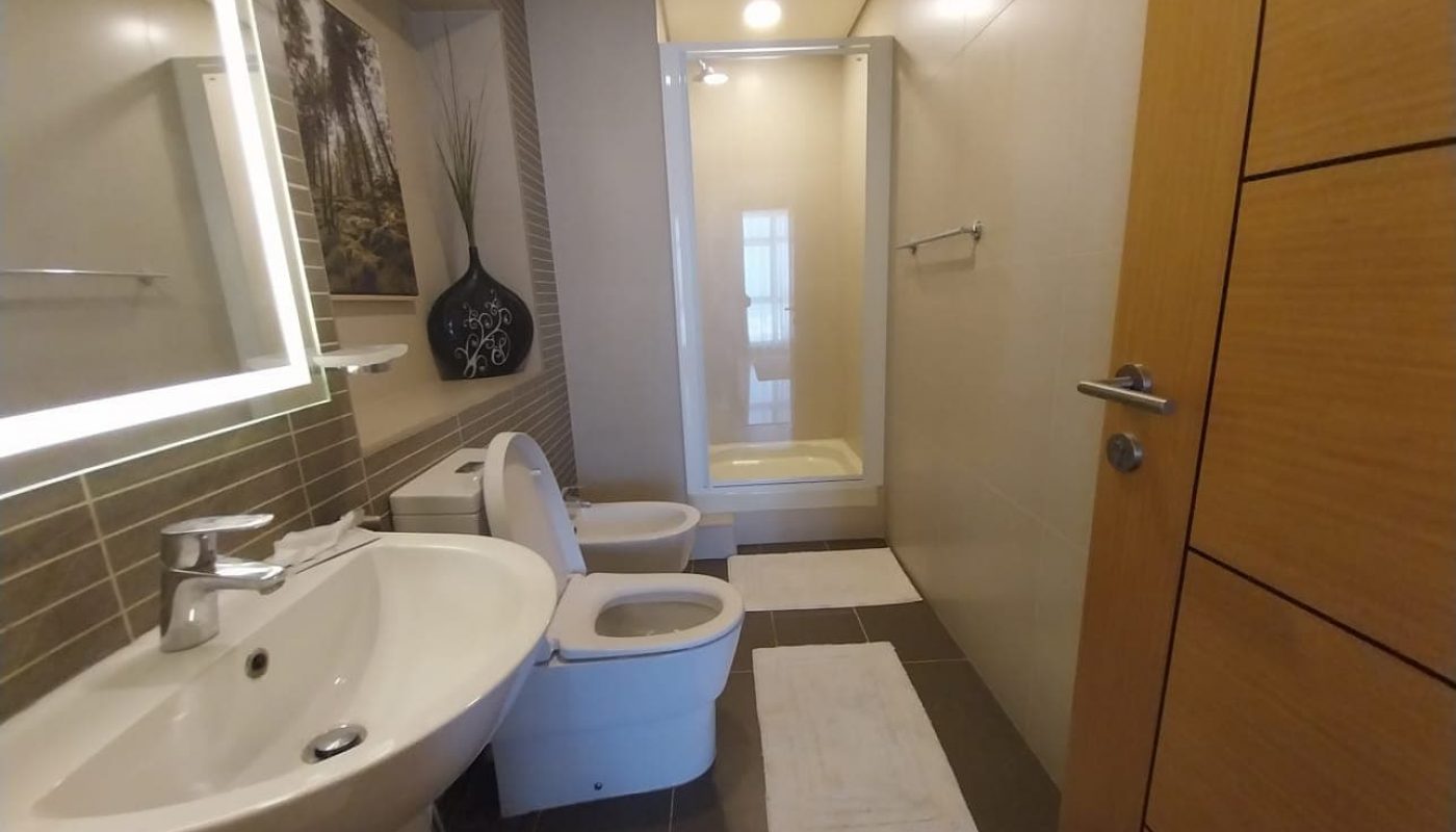Modern bathroom with a sink, toilet, bidet, towel rack, and a shower stall at the far end, featuring neutral-colored tiles and a wooden door.