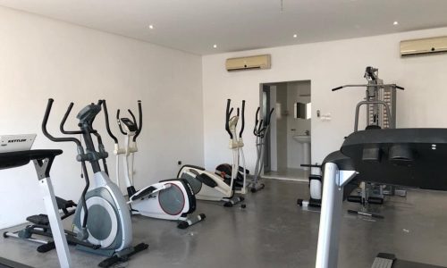 A small, modern home gym equipped with treadmills, elliptical machines, and a weight training station, featuring a minimalist white decor.