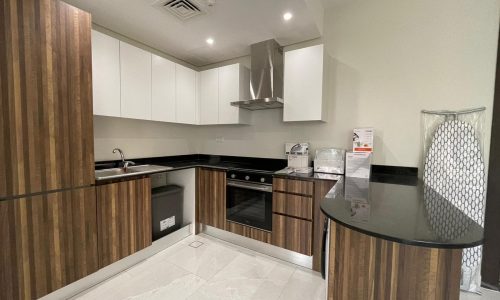 A modern kitchen in this AMWAJ flat for sale features dark wood cabinetry, black countertops, stainless steel appliances, a large white floor tile, and a freestanding ironing board.