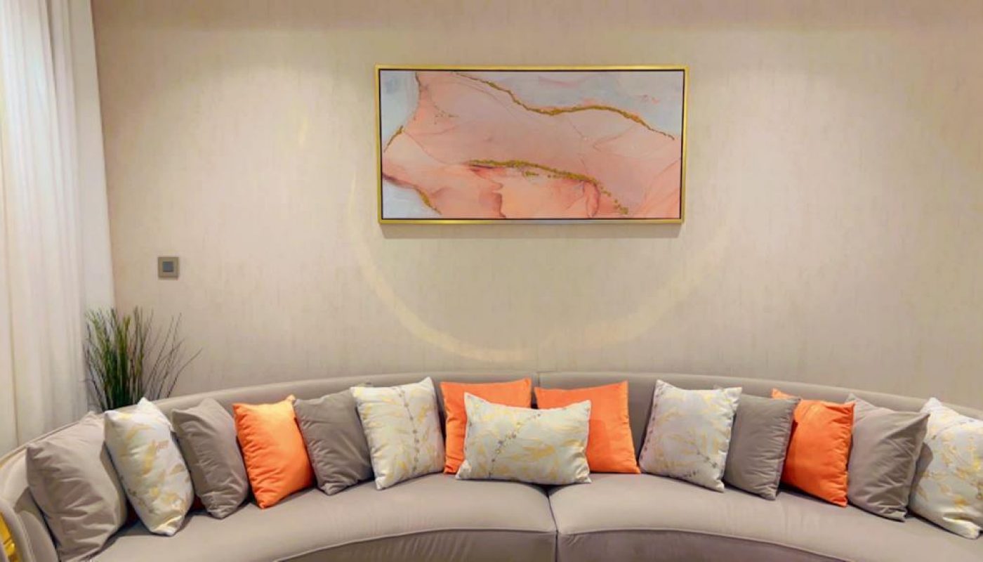 A semi-circular gray sofa with orange, white, and gray cushions is in front of a beige wall with a framed abstract painting. A small plant is positioned to the left.