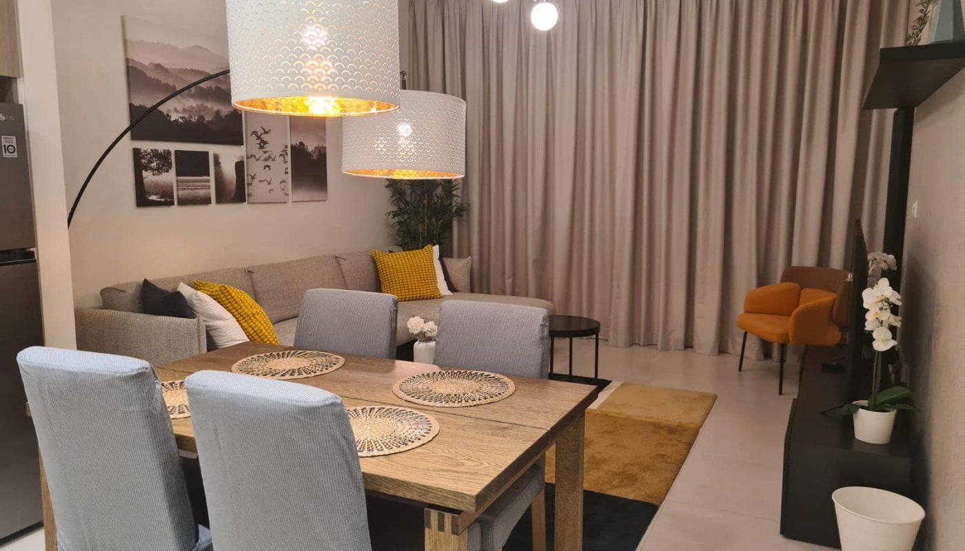A modern living room and dining area within a flat for sale in Diyar Al Muharaq features a wooden table, gray chairs, a gray sofa, an orange accent chair, white curtains, and contemporary lighting fixtures. Decorative elements and artwork enhance the space.