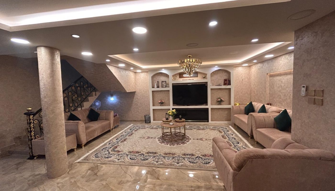 A spacious living room with beige sofas, a patterned rug, built-in shelves, a large TV, decorative lighting, and marble flooring.