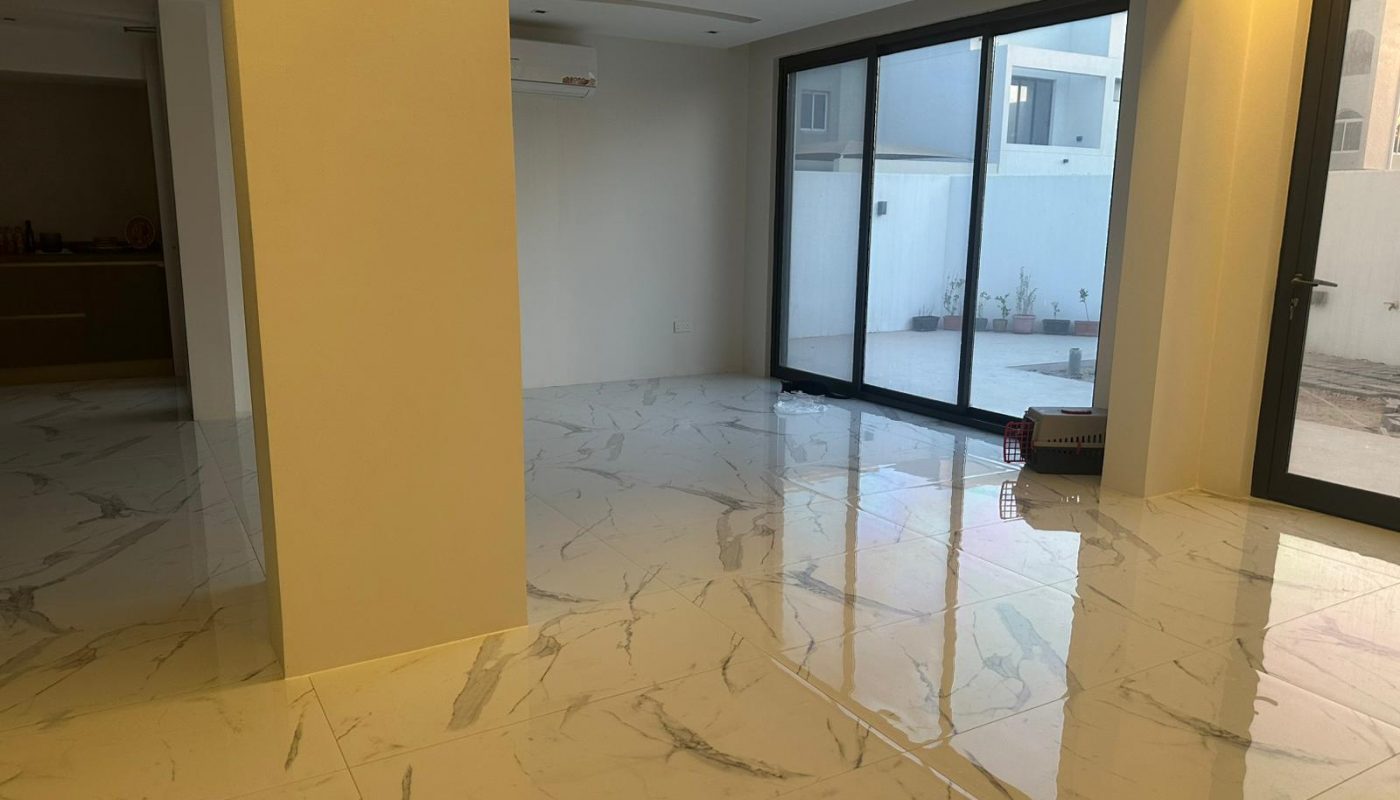 Spacious modern room with white marble floors, large glass sliding doors, and minimal furniture. Outdoor area visible through the windows.