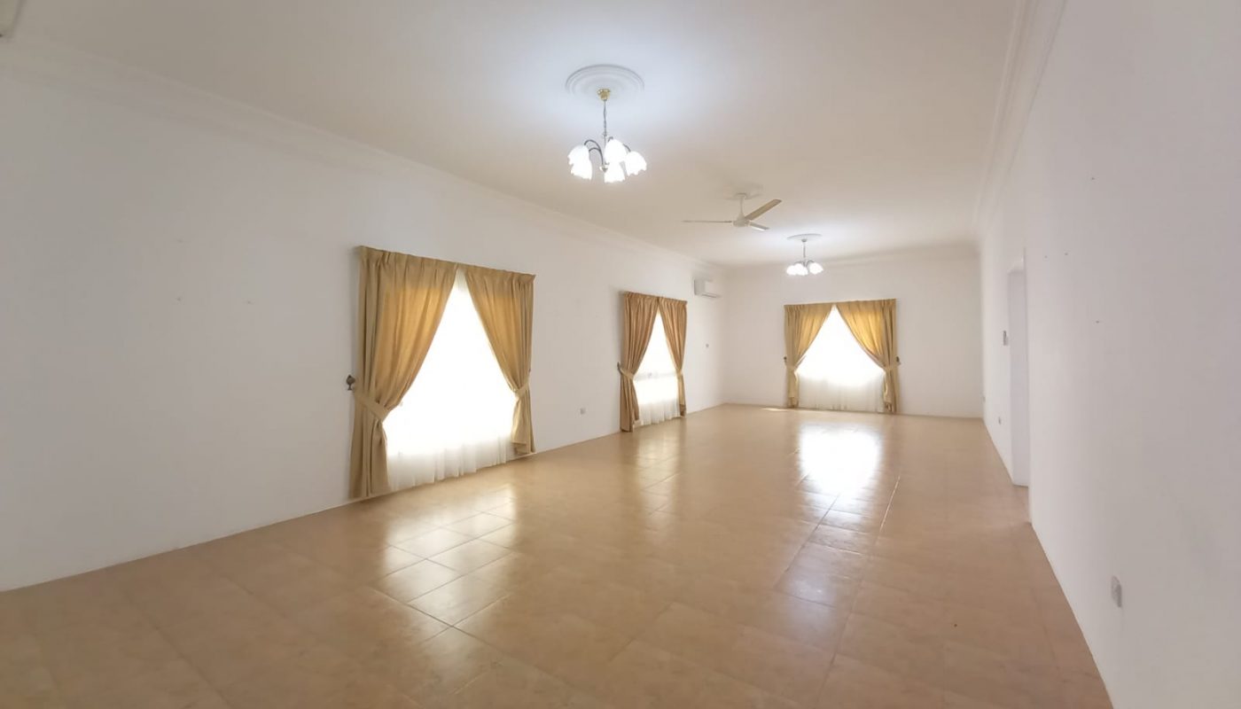 Empty, brightly lit room with white walls, tiled floor, three windows with gold curtains, and two ceiling fans.