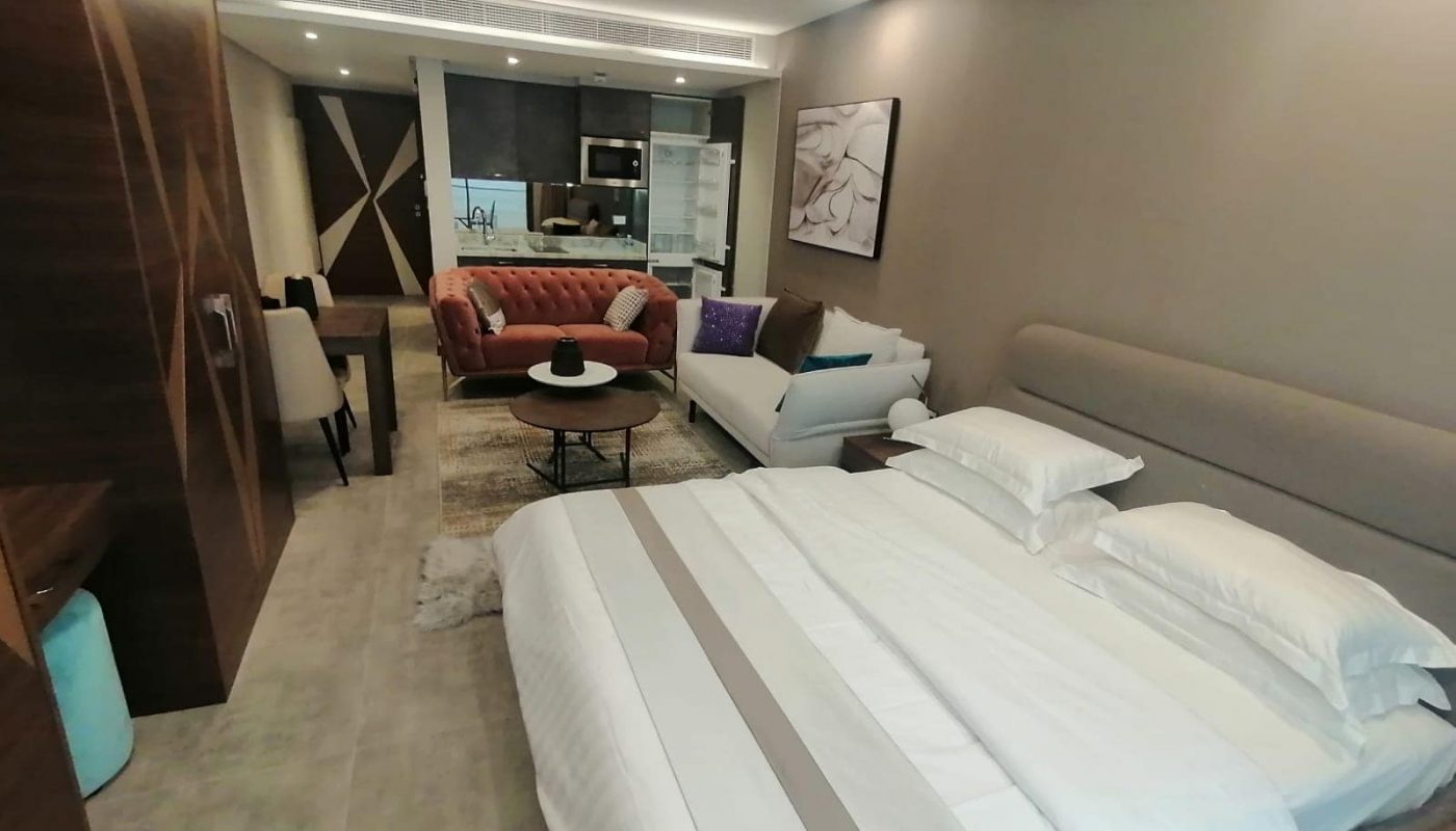 Modern studio apartment with a neatly made bed, two sofas, a coffee table, and a kitchenette in the background. Neutral color palette with wooden accents and contemporary decor.