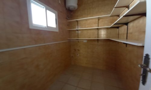 Interior of an empty storage room with beige tiled walls, multiple white shelves, and a small window.