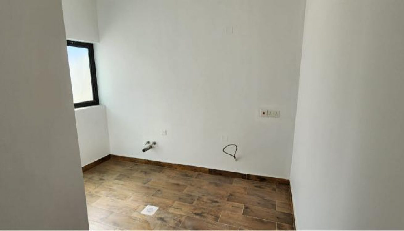 Empty room with white walls, a small window on the left, brown wood-look floor tiles, and exposed plumbing and electrical outlets on one wall.